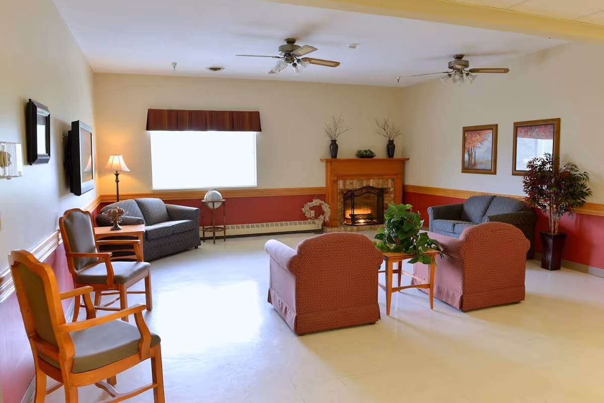 A cozy living room area with a fireplace, two ceiling fans, and a window with a valance. The room features two armchairs and a loveseat arranged around a small wooden table with a plant. There are additional chairs along the wall, framed artwork, a floor lamp, and decorative plants.