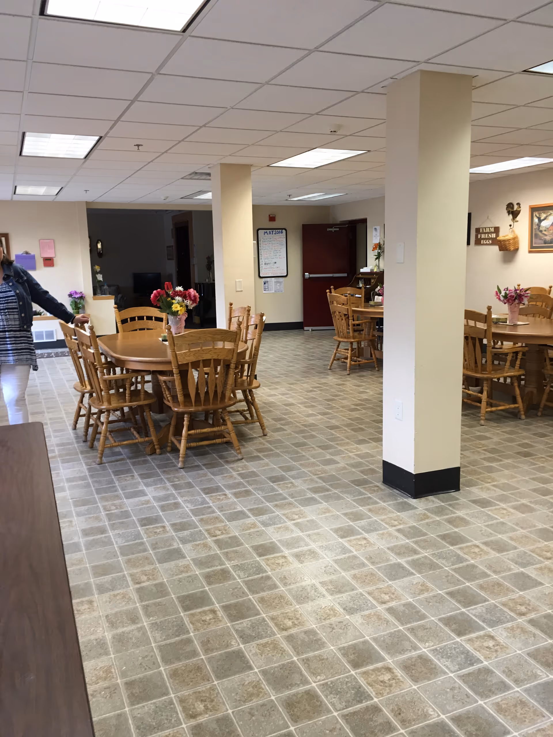 Interior view of a dining area with multiple wooden tables and chairs arranged on a tiled floor. Each table has a vase with colorful flowers. There are two large support columns in the room, and a person partially visible on the left side. The walls have some decorations including a sign that reads 'WARM FRESH EGGS' and a framed picture. A red door and a calendar are visible in the background.