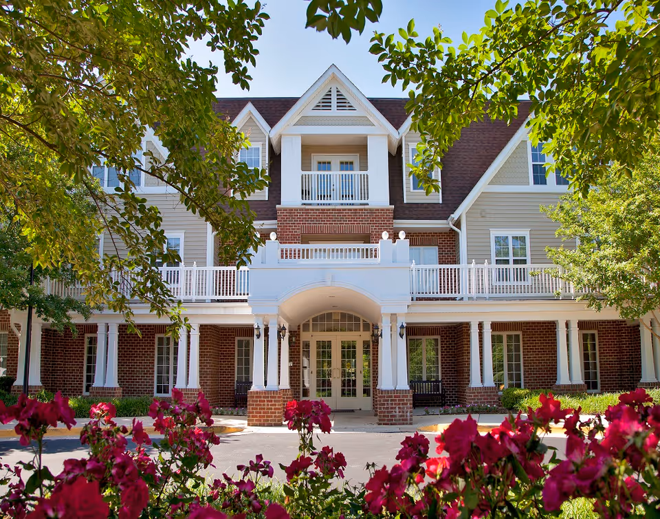 Front entrance of a multi-story brick and siding senior living building with columns, balconies, and flowers in the foreground.
