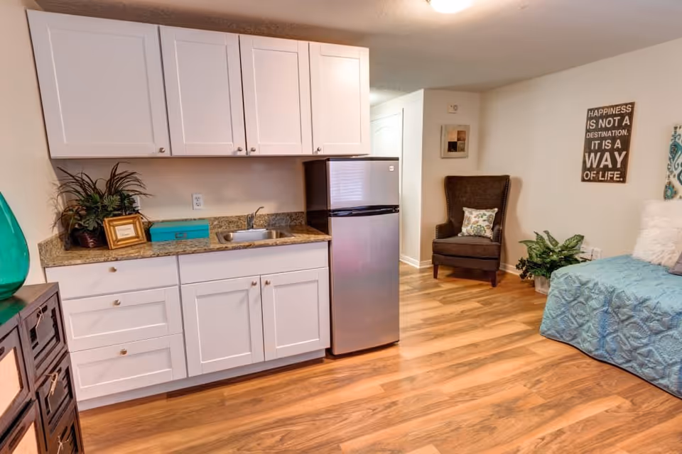A small kitchenette area with white cabinets, a granite countertop, a small sink, and a stainless steel mini refrigerator. To the right, there is a cozy corner with a dark brown armchair, a green plant, and a blue quilted sofa with pillows. The room has wood flooring and light-colored walls with a decorative sign that reads 'Happiness is not a destination. It is a way of life.'
