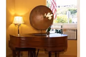 A wooden grand piano with a decorative round metal plate and a small flower arrangement on top, positioned near a window with an air conditioning unit below it. A table lamp with a beige shade is placed on the left side of the piano, illuminating the warm-toned room.