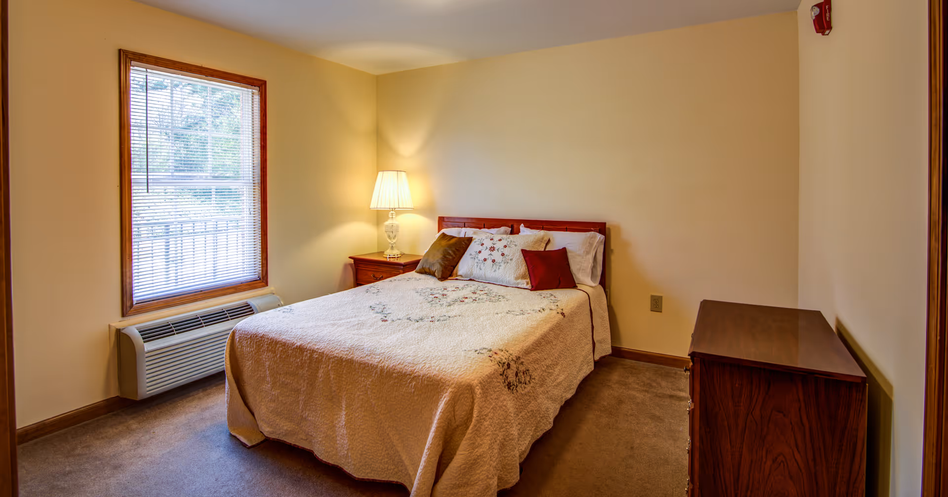A cozy bedroom with a double bed covered in a beige quilt with floral embroidery. There are several pillows on the bed, including two red ones. A wooden nightstand with a lamp is positioned next to the bed. A window with blinds is on the left wall, and a wooden dresser is on the right side of the room. The walls are painted light yellow, and the carpet is brown.