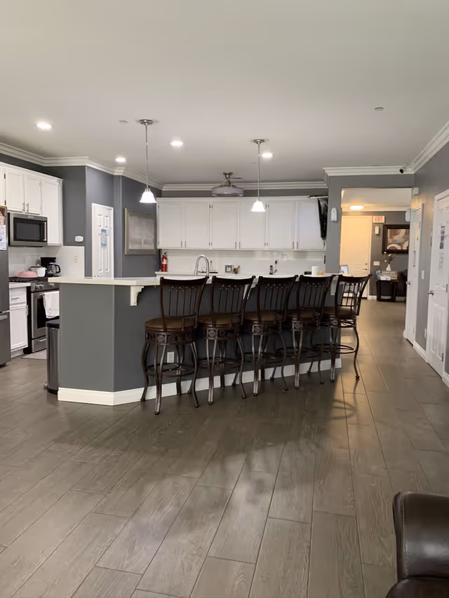 A modern kitchen area with white cabinets, a gray island with a white countertop, and five high chairs lined up along the island. The kitchen features stainless steel appliances including a microwave and stove, pendant lights hanging above the island, and a ceiling fan. The floor is covered with wood-like tiles, and there is a hallway visible in the background.