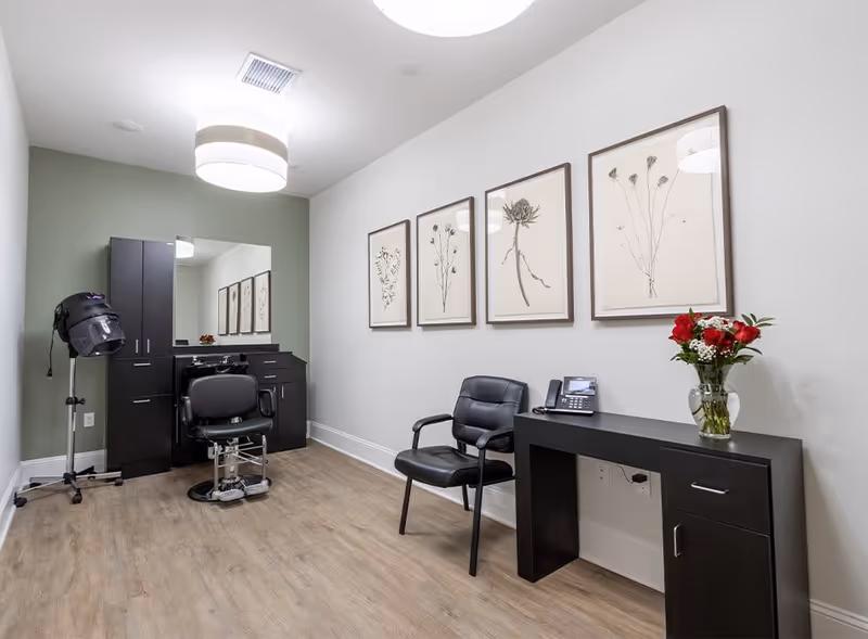 A modern salon room with a black salon chair in front of a mirror and cabinetry, a hair dryer on a stand, a black chair next to a black desk with a telephone and a vase of red and white flowers, and four framed botanical prints on the wall.