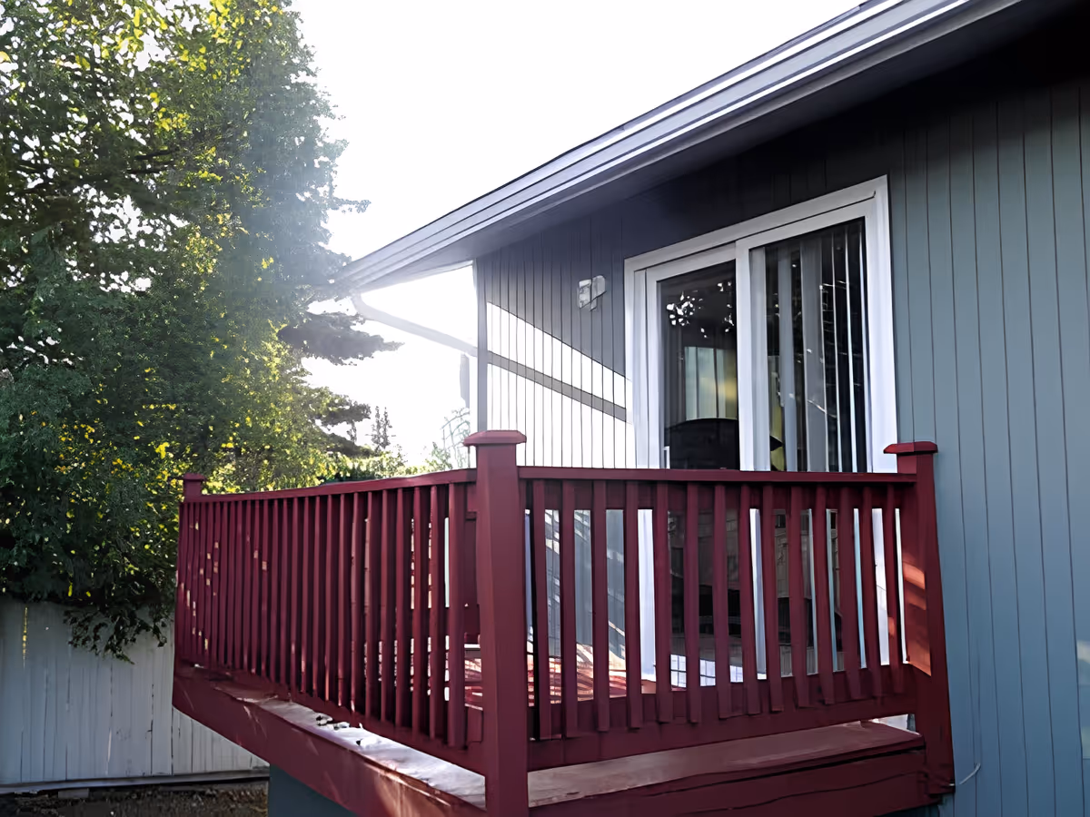 Small exterior deck with red railing attached to a light-blue house and sliding glass door, with trees and a white fence in the background.