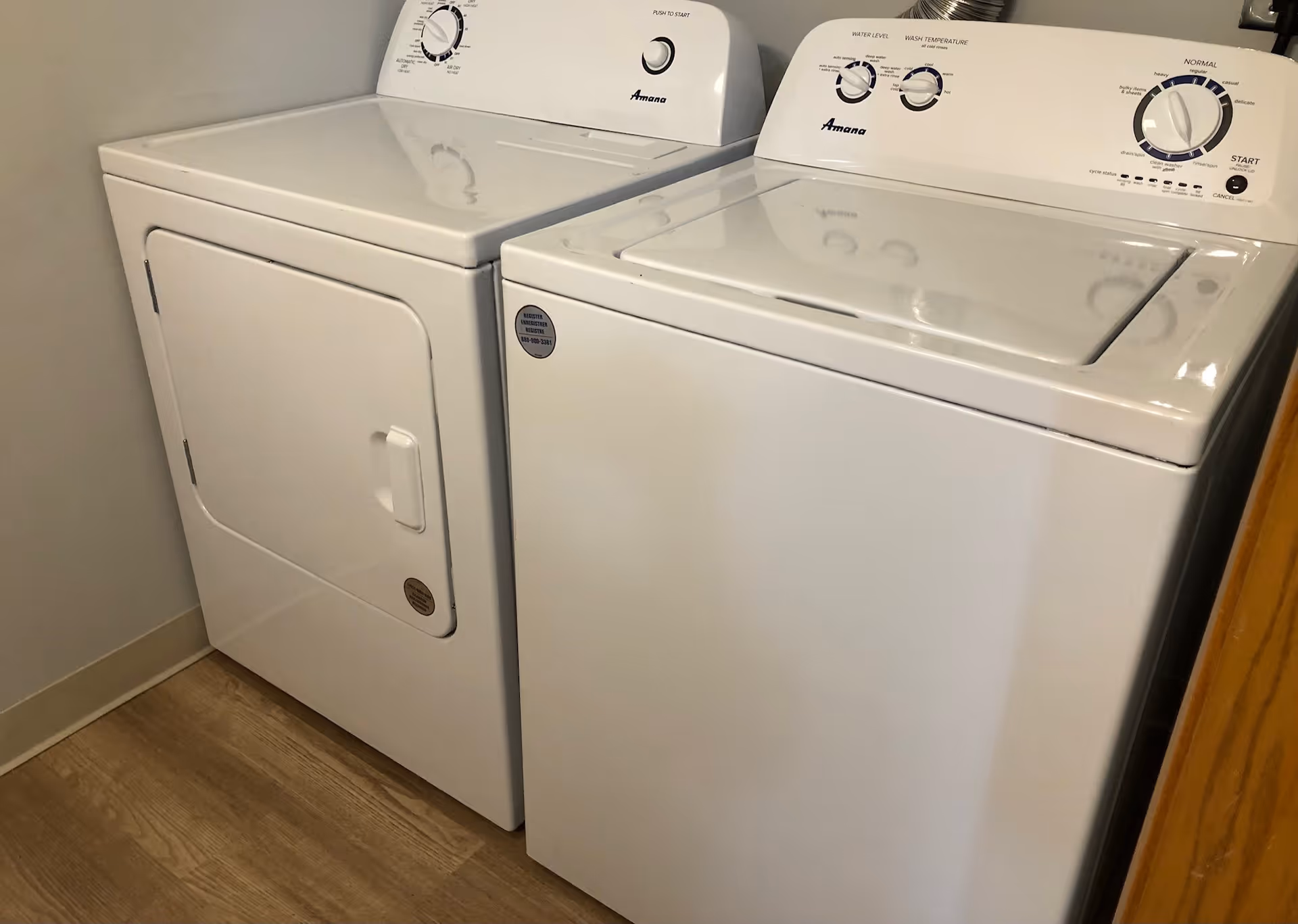 A white washing machine and dryer set placed side by side in a laundry area with light-colored wooden flooring and beige walls.