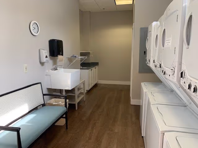 Laundry room with multiple stacked white washers and dryers on the right side, a bench with a light blue cushion on the left, a utility sink, and cabinets along the back wall. The room has wood flooring and neutral-colored walls with a clock and soap dispensers mounted on the wall.