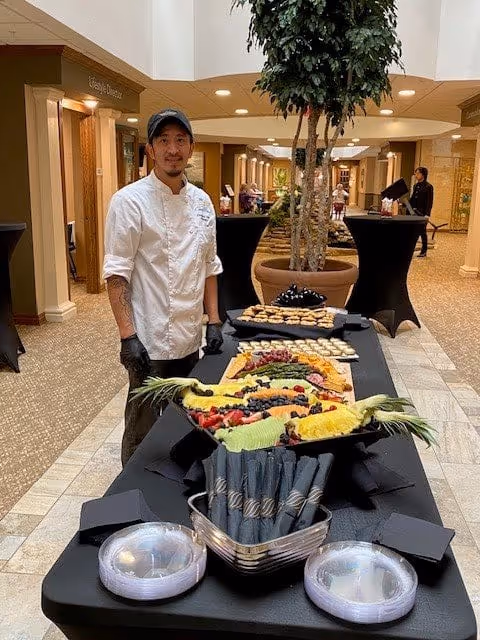 A chef wearing a white coat and black gloves stands behind a long table covered with a black tablecloth. The table displays an assortment of food, including a large fruit platter with various sliced fruits and berries, and trays of crackers and cheese. The setting appears to be an indoor common area with potted plants and people in the background.
