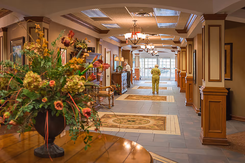 A warmly decorated retirement community lobby with a large flower arrangement in the foreground and residents walking down the hallway toward the windows.