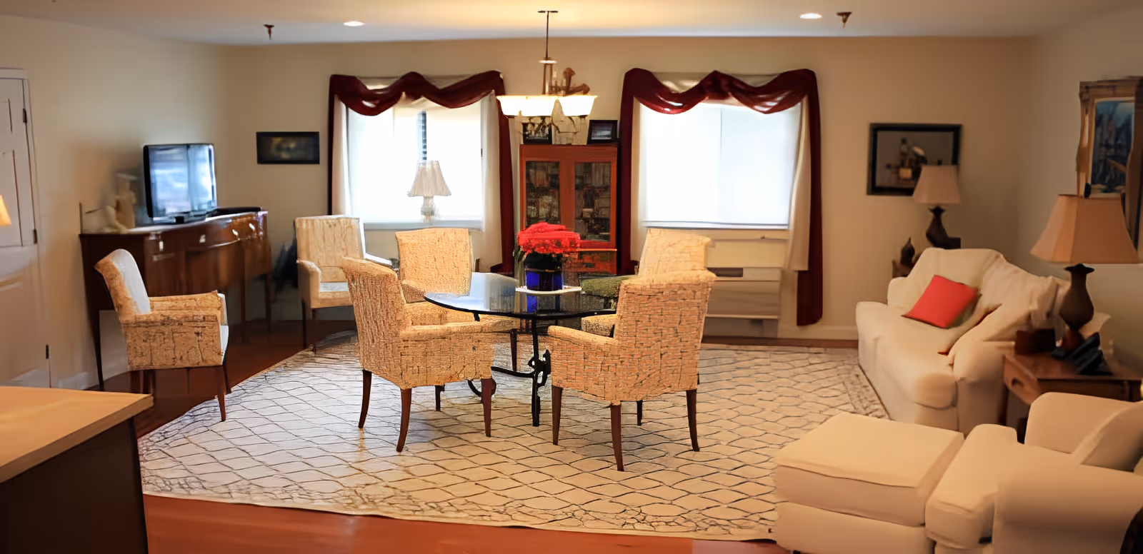 A cozy living room area with a glass-top round table surrounded by six upholstered chairs on a patterned rug. There is a white sofa with a red pillow, a white armchair with an ottoman, two windows with curtains, a wooden cabinet, a TV on a sideboard, and several lamps providing warm lighting.