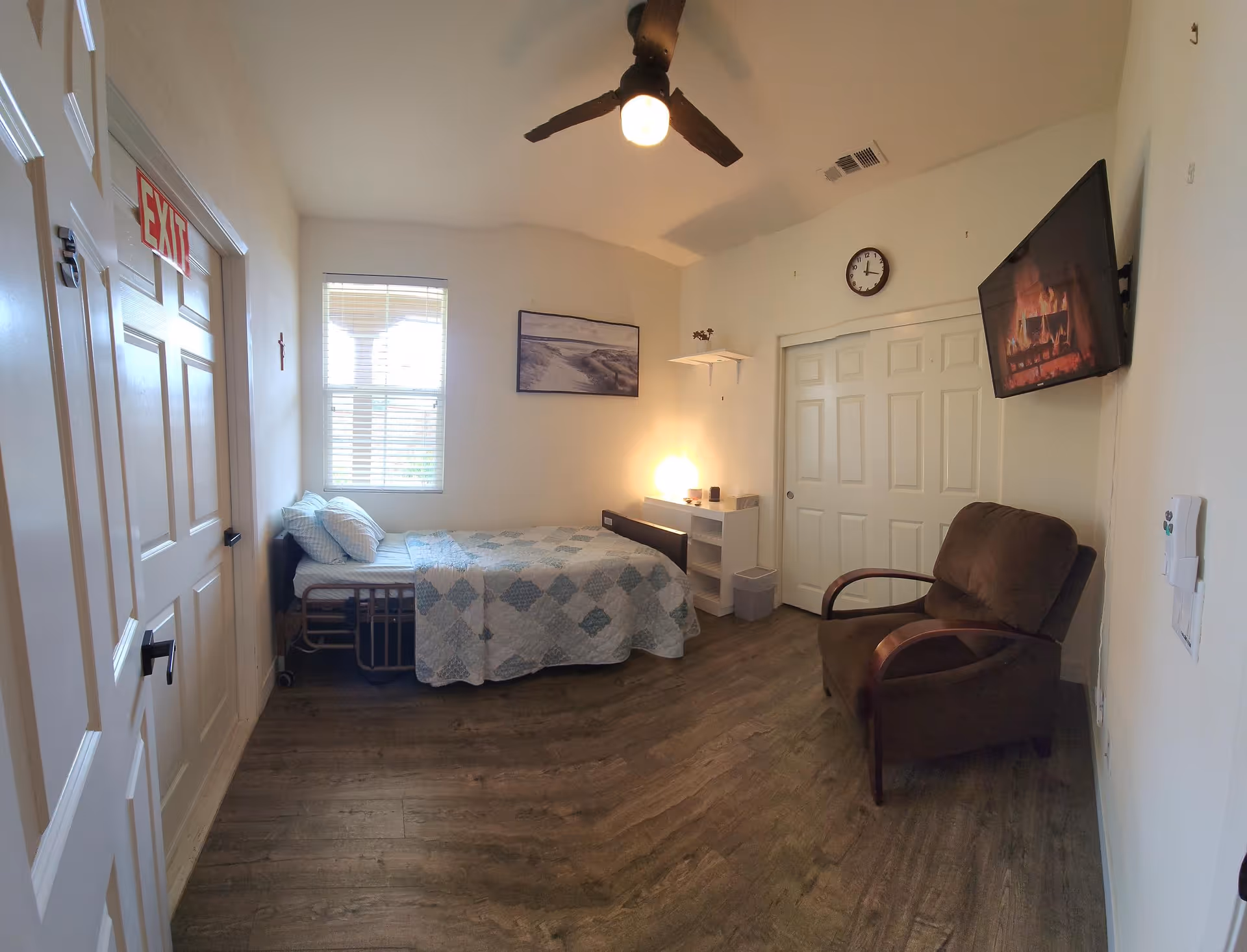 A small, cozy bedroom with a single bed covered in a blue and white quilt, a brown armchair, a wall-mounted TV displaying a fireplace, a ceiling fan with a light, a window with blinds, a clock on the wall, and a white closet with sliding doors.