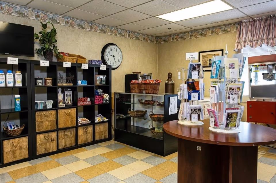 Interior view of a small shop area in a senior living facility with shelves holding personal care items, decorative objects, and baskets. A clock is mounted on the wall above the shelves. A glass display case contains snacks, and a round table displays greeting cards on rotating racks. The room has a tiled floor and a window with a valance.