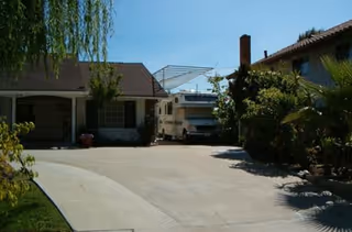 Driveway area between two residential-style buildings with a parked RV under a metal carport structure. Trees and shrubs are visible around the buildings under a clear blue sky.