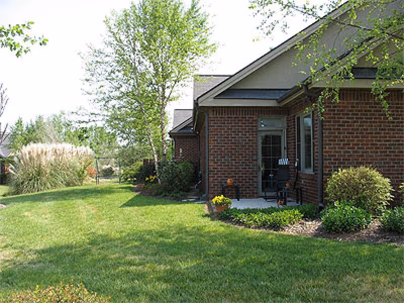 Brick building exterior with a small patio, lawn, and landscaped shrubs and trees.