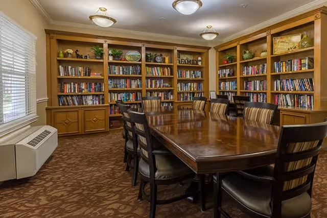 A well-lit room with a large wooden table surrounded by chairs with striped cushions. The walls are lined with wooden bookshelves filled with books and decorative items. A window with blinds is on the left side, and the floor is carpeted with a patterned design.