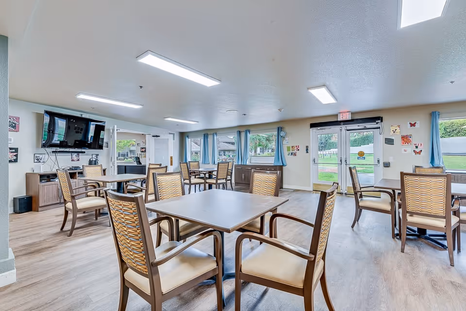 A bright and spacious common dining area in a senior living facility with multiple square tables surrounded by wooden chairs with cushioned seats. The room has large windows with blue curtains letting in natural light, a wall-mounted TV, and double glass doors leading outside. The walls are decorated with small colorful artworks.