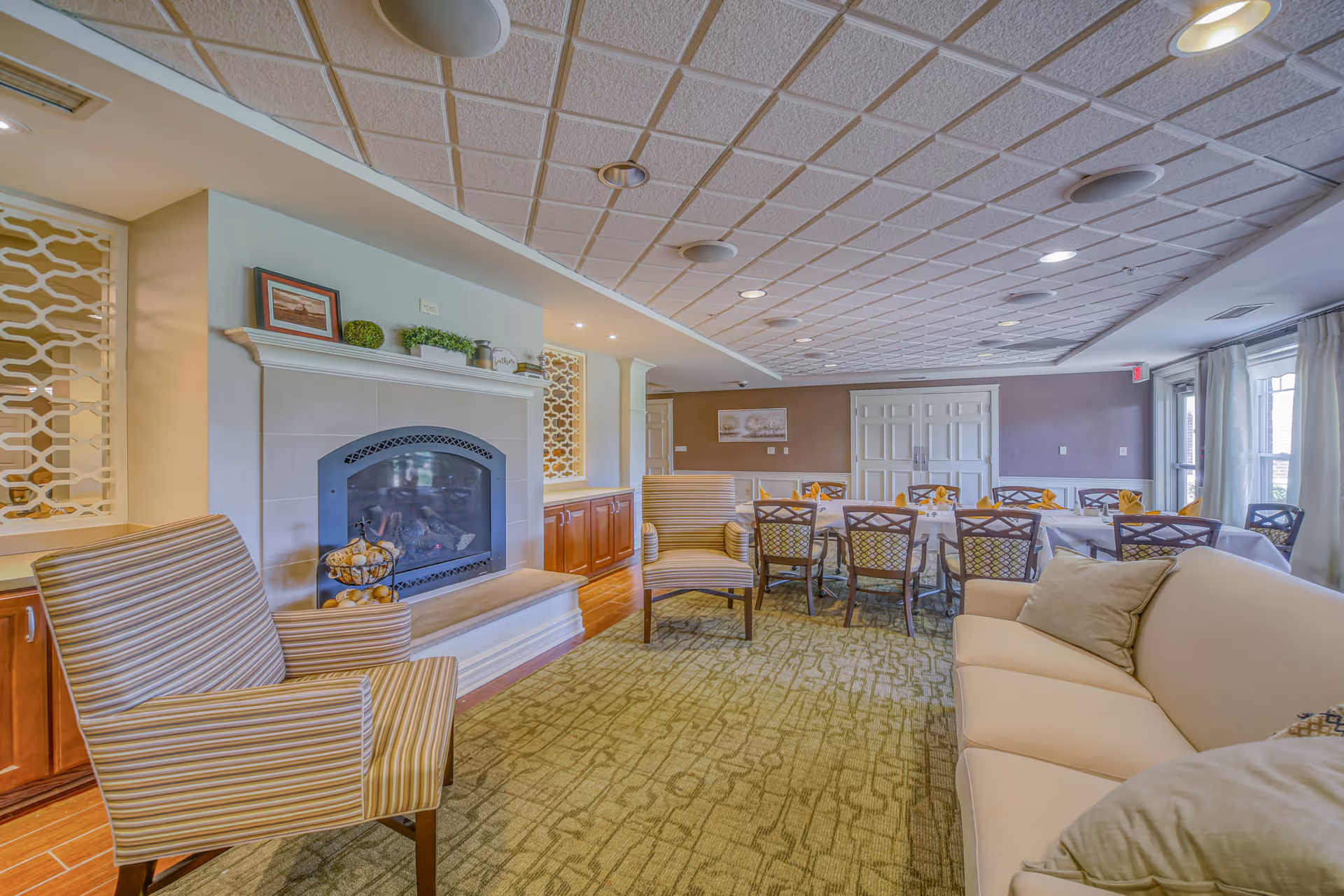 A cozy assisted living common area featuring a fireplace with decorative items on the mantel, two striped armchairs, a beige sofa with cushions, and a dining area with tables and chairs set with napkins. The room has a patterned carpet, a tiled ceiling with recessed lighting, and large windows with curtains allowing natural light.