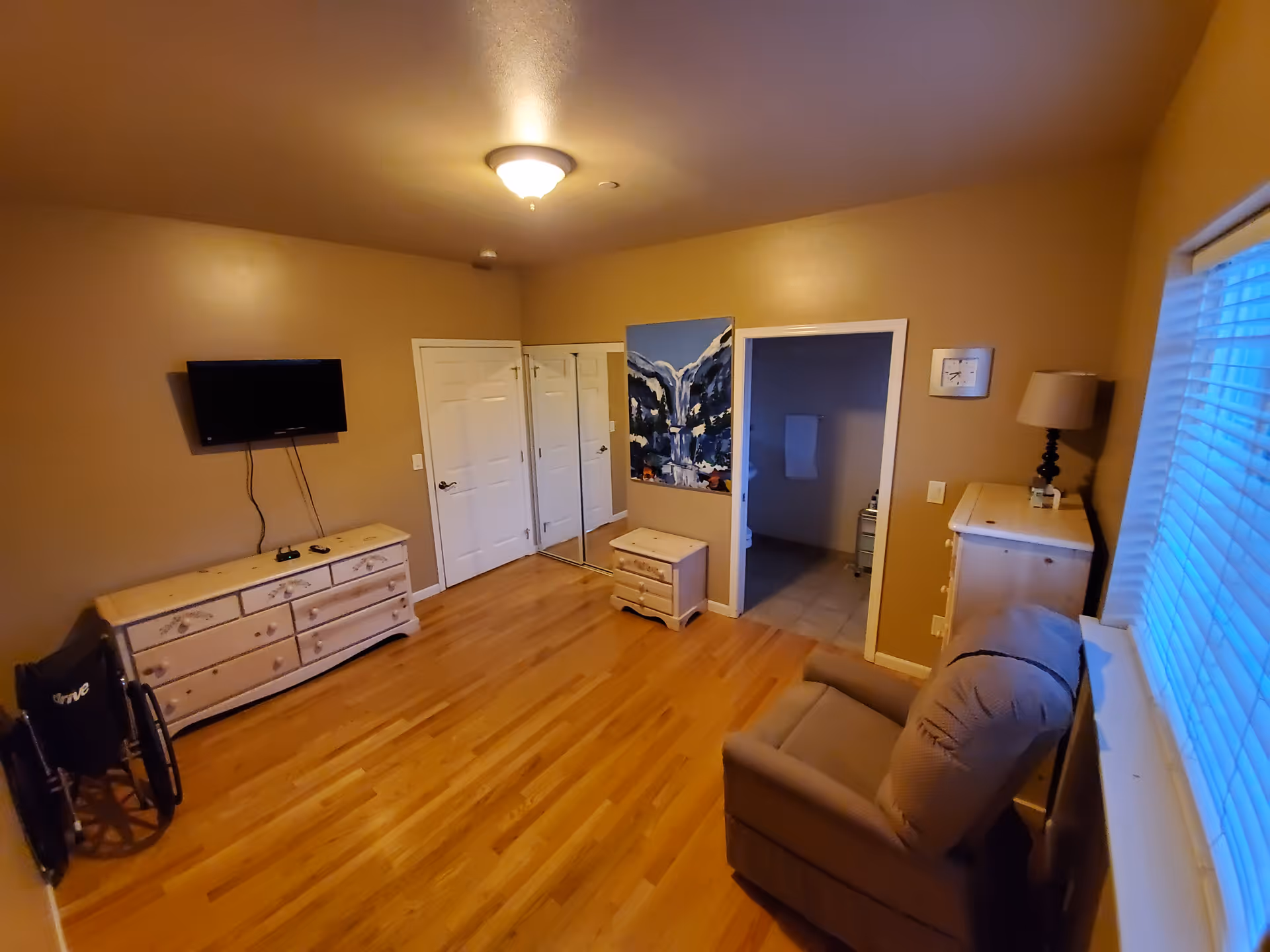 A room with wooden flooring and beige walls featuring a mounted flat-screen TV above a white dresser, a wheelchair, a small white nightstand, a beige armchair near a window with blinds, a lamp on a white chest of drawers, and a doorway leading to a bathroom with tiled floor and a towel hanging on the wall.