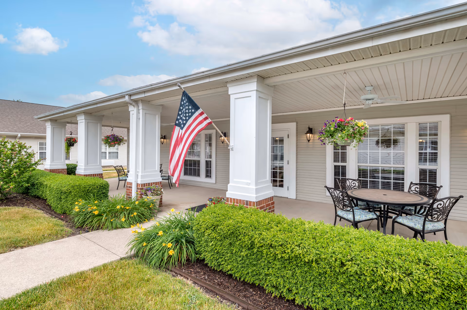 Covered outdoor patio area with white columns, hanging flower baskets, an American flag, a round table with four chairs, and well-maintained green bushes and yellow flowers along the walkway.