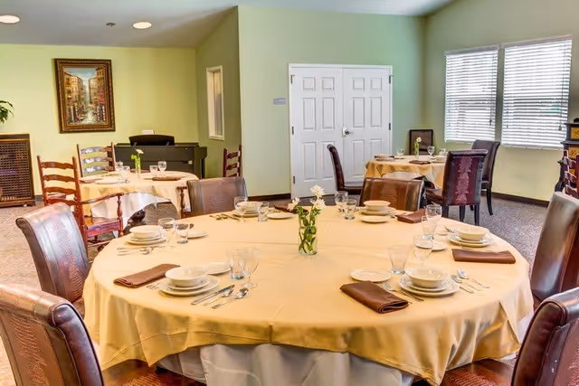 A dining room with round tables covered in beige tablecloths, each set with plates, bowls, glasses, silverware, and brown napkins. There are several chairs around each table, and a small vase with flowers is placed at the center of the main table. The room has light green walls, a window with blinds, a painting on the wall, and a double door in the background.