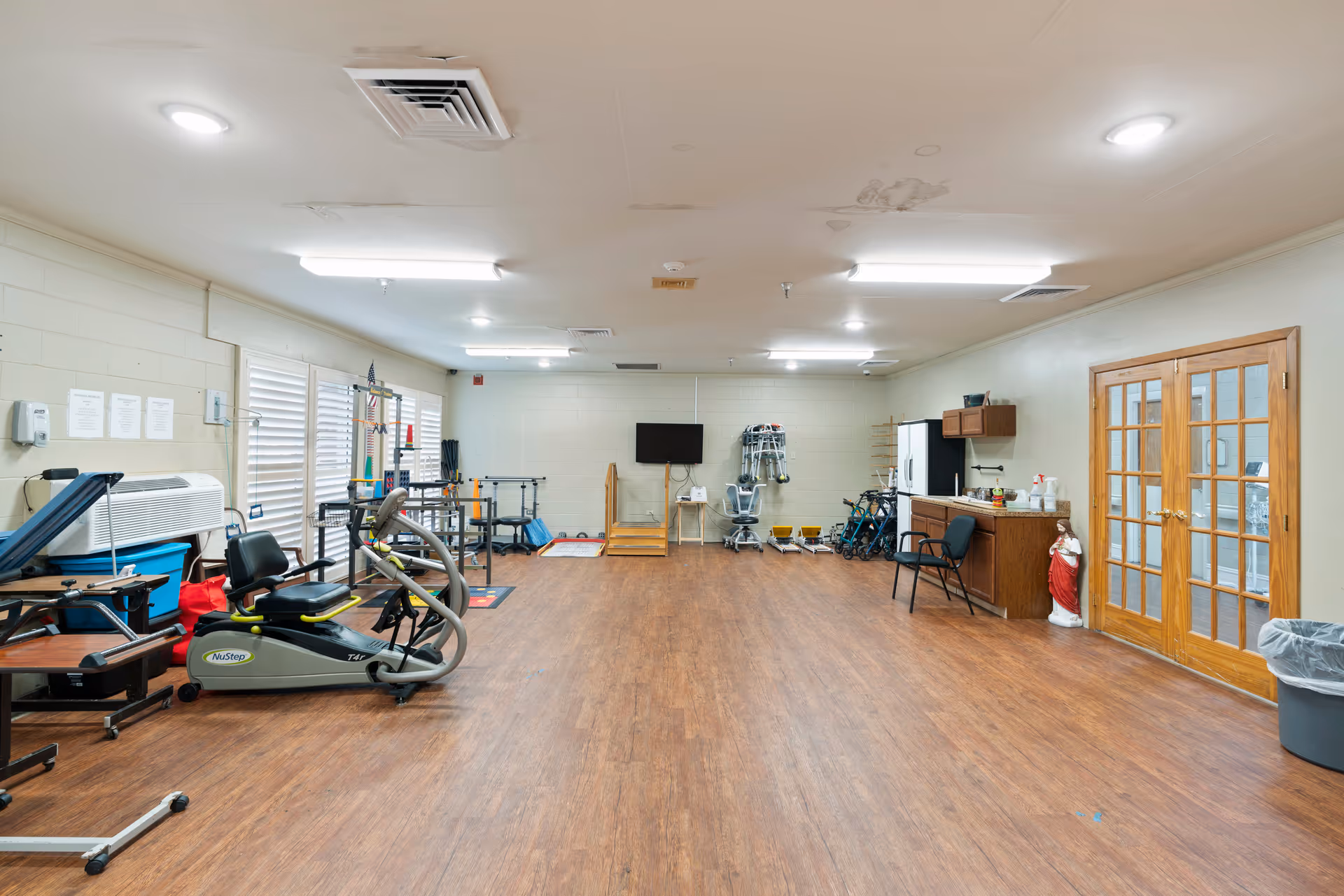 A spacious rehabilitation or therapy room with wooden flooring, exercise equipment including a NuStep machine, parallel bars, and various mobility aids. The room has white walls, fluorescent ceiling lights, a wall-mounted TV, a small kitchenette area with a chair, and double wooden doors with glass panels. There is also a statue of a religious figure near the kitchenette.