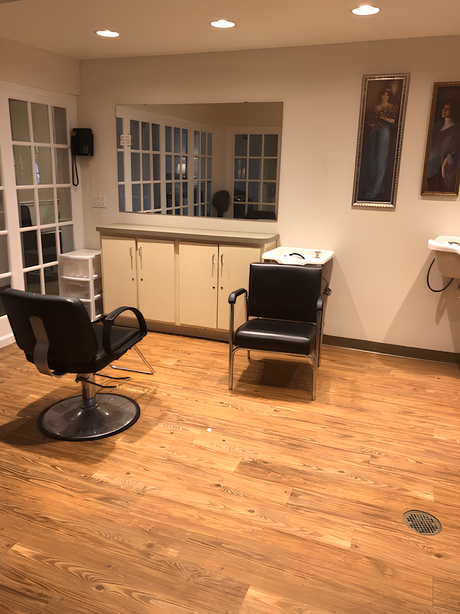Interior of a salon area with two black salon chairs, a large mirror on the wall, a cabinet with drawers, two sinks for hair washing, wooden flooring, and two framed paintings on the wall.