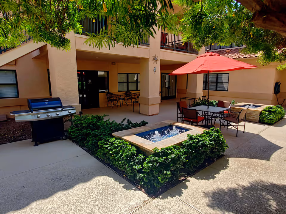 Outdoor courtyard area at Oasis at Mesa Palms featuring a small water fountain surrounded by greenery, a patio table with chairs and a red umbrella, a barbecue grill, and shaded seating areas under the building overhang.
