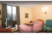 Single-occupancy bedroom with a bed covered in a pink blanket, an overbed table, a green chair, a TV on a dresser, and a large window with curtains.