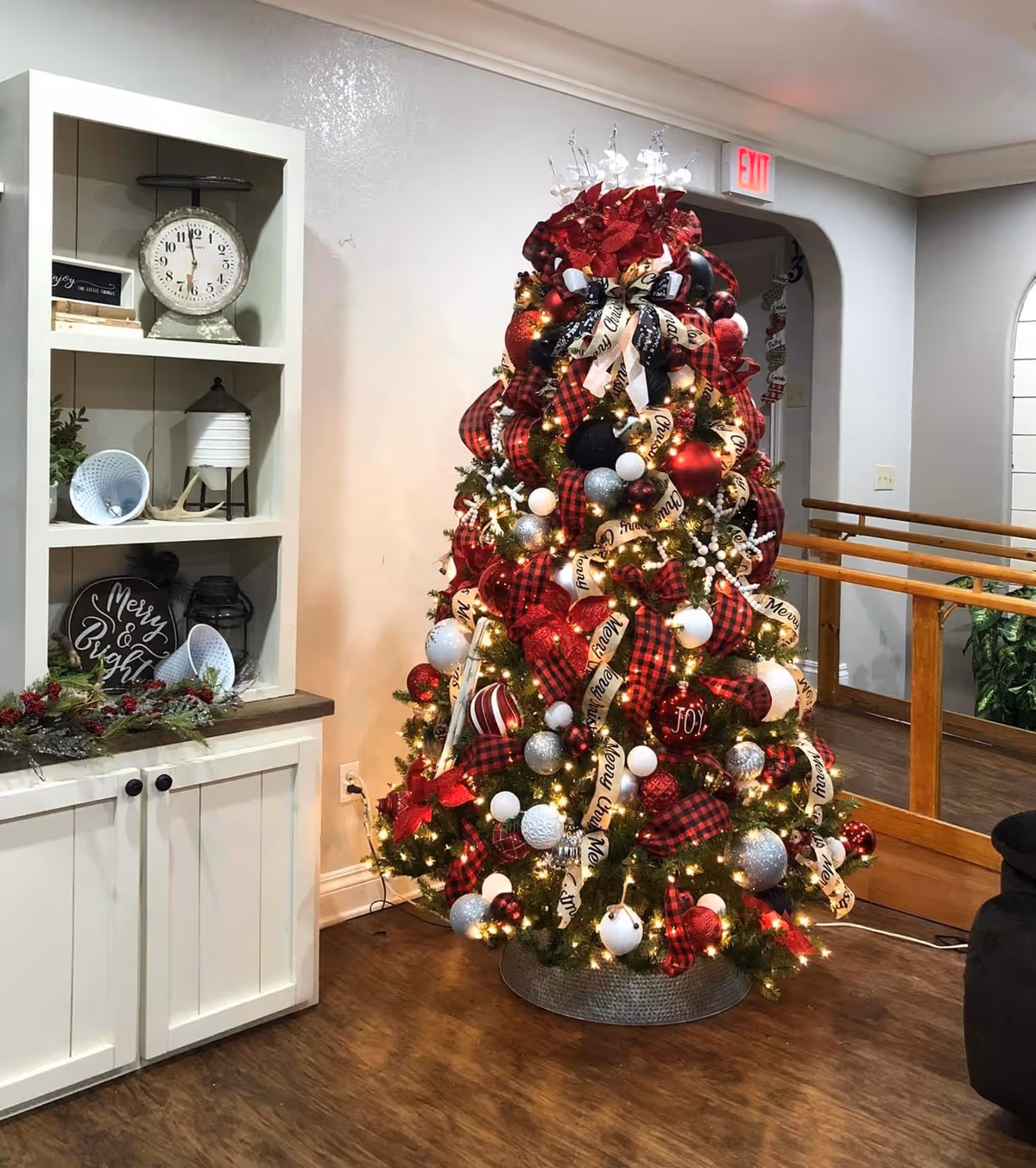 Festively decorated Christmas tree with red plaid ribbons and ornaments in a residential living area beside a white shelf.