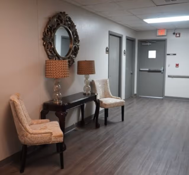 A hallway in an assisted living facility with two upholstered chairs and a dark wooden console table between them. On the table are two matching lamps with textured shades, and above the table is a decorative round mirror with an ornate frame. The hallway has gray doors and a gray floor, with an exit door at the end.