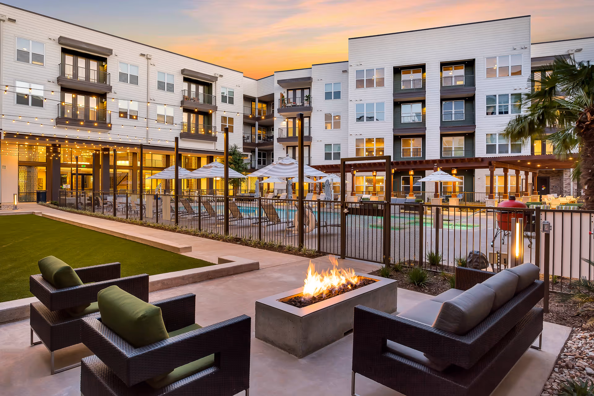 Outdoor seating area with modern wicker chairs and a rectangular fire pit in the foreground, overlooking a fenced swimming pool with lounge chairs and umbrellas. The background shows a multi-story residential building with balconies and warm interior lighting at sunset.