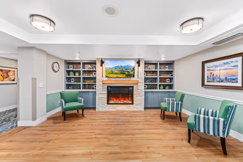 A bright communal lounge featuring an electric fireplace with a mounted TV, built-in shelving, and green upholstered chairs.