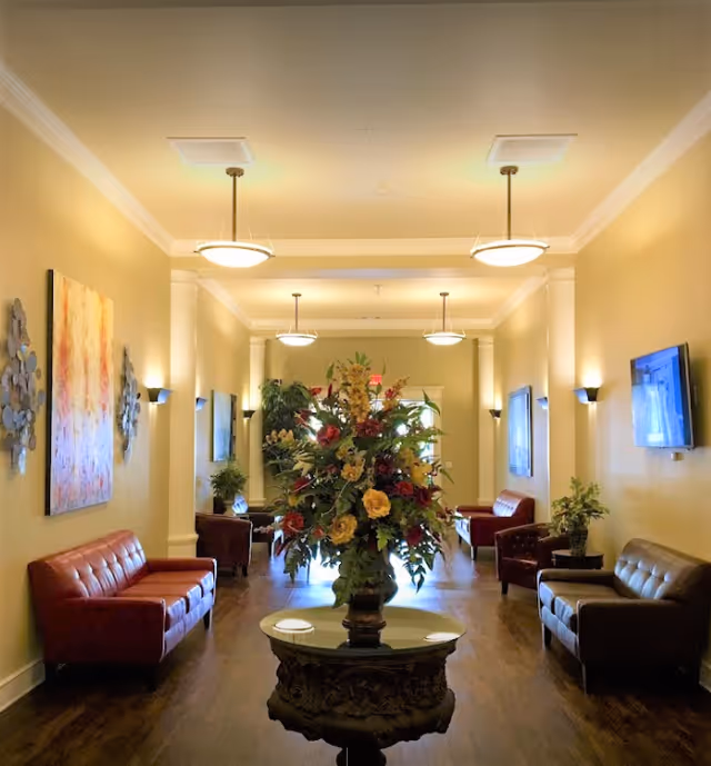 A well-lit lounge area with several brown leather sofas arranged along the walls. A large floral arrangement sits on an ornate round table in the center of the room. The walls are painted a warm beige color and decorated with artwork and wall sconces. Ceiling lights hang evenly spaced along the length of the room.
