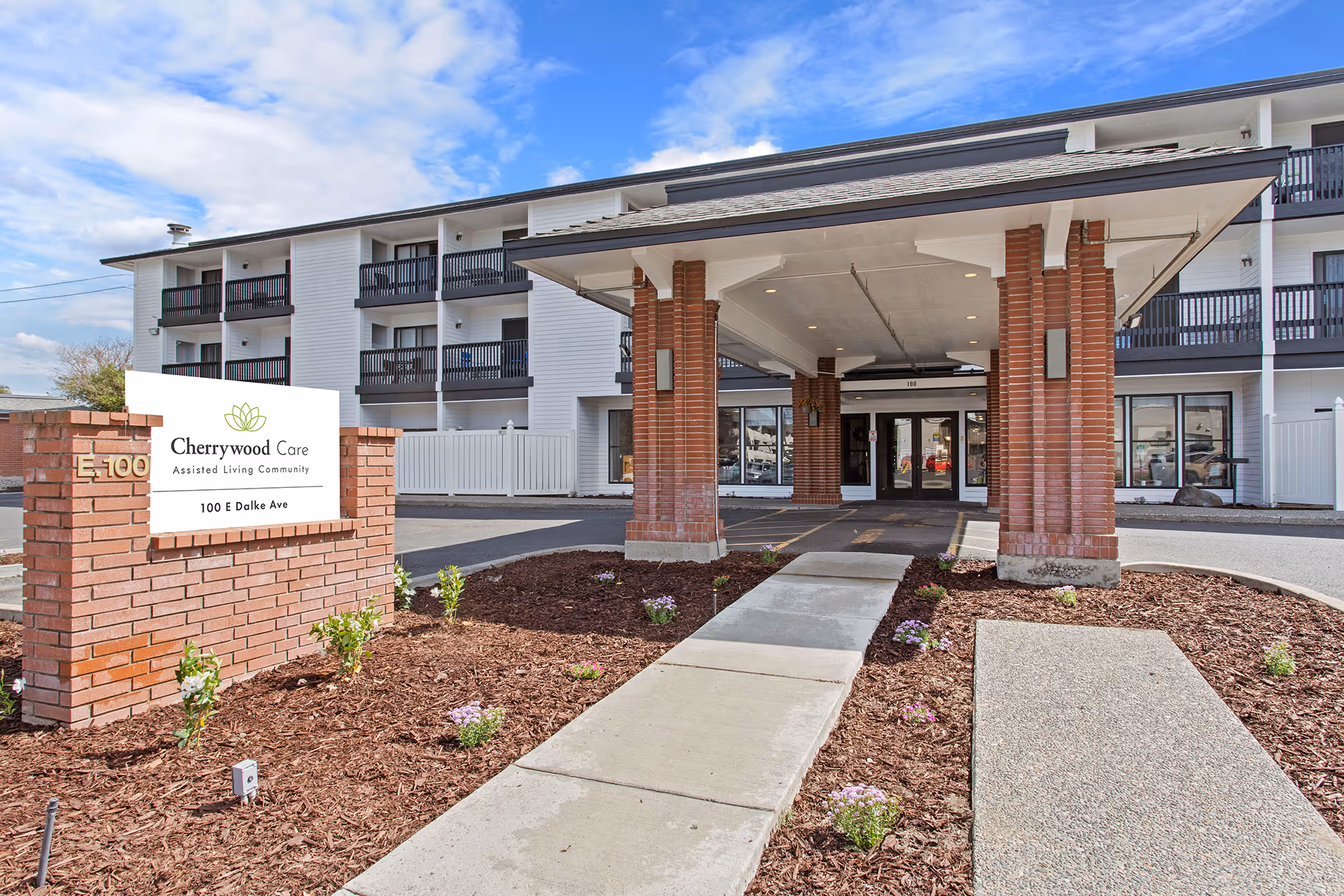 Entrance of Cherrywood Care Assisted Living Community showing a covered driveway with brick pillars, a sidewalk leading to the main doors, and a sign with the facility name and address 100 E Dalke Ave. The building is white with black balconies and a clear blue sky above.