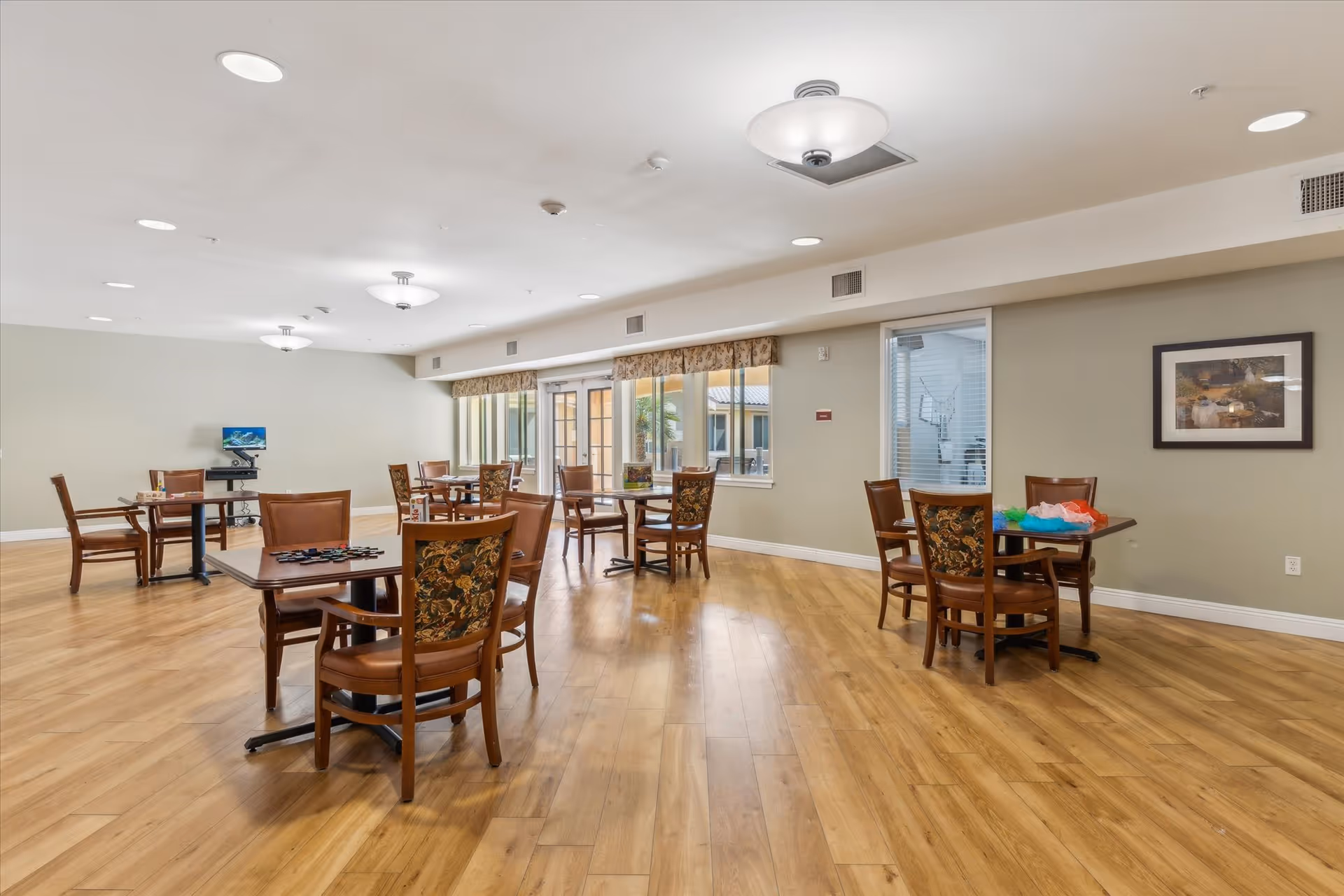 A spacious room with wooden flooring and several small tables surrounded by chairs. The room has large windows with valances, allowing natural light to enter. The walls are painted light green, and there is a framed picture hanging on one wall. The ceiling has multiple light fixtures, and there is a computer station in the back corner.