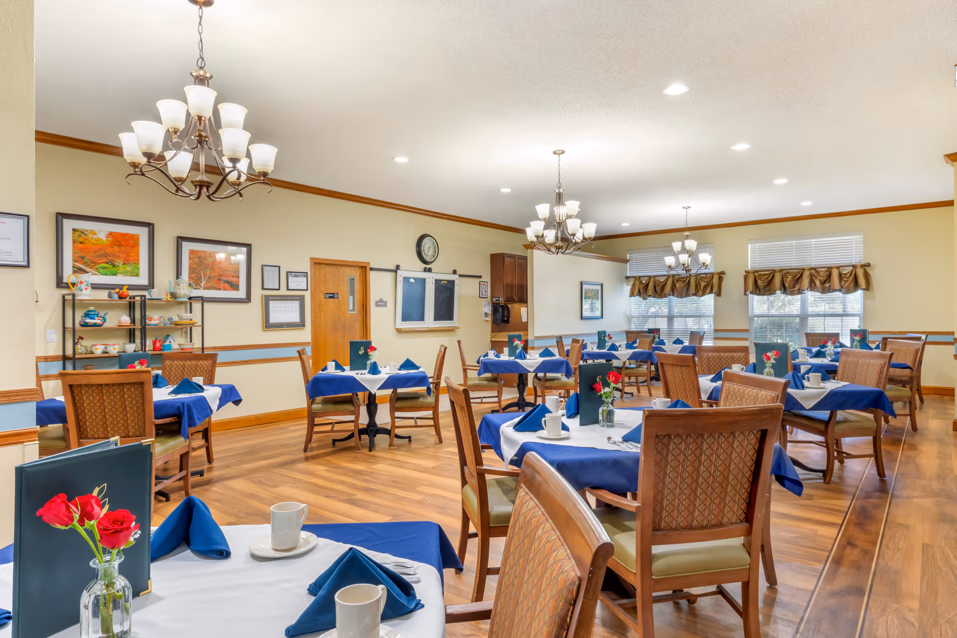 A dining room in a senior living facility with multiple tables covered in white and blue tablecloths, each set with cups, napkins, and small vases holding red flowers. The room has wooden chairs, hardwood floors, beige walls with blue trim, and several chandeliers hanging from the ceiling. There are windows with valances letting in natural light and framed pictures on the walls.