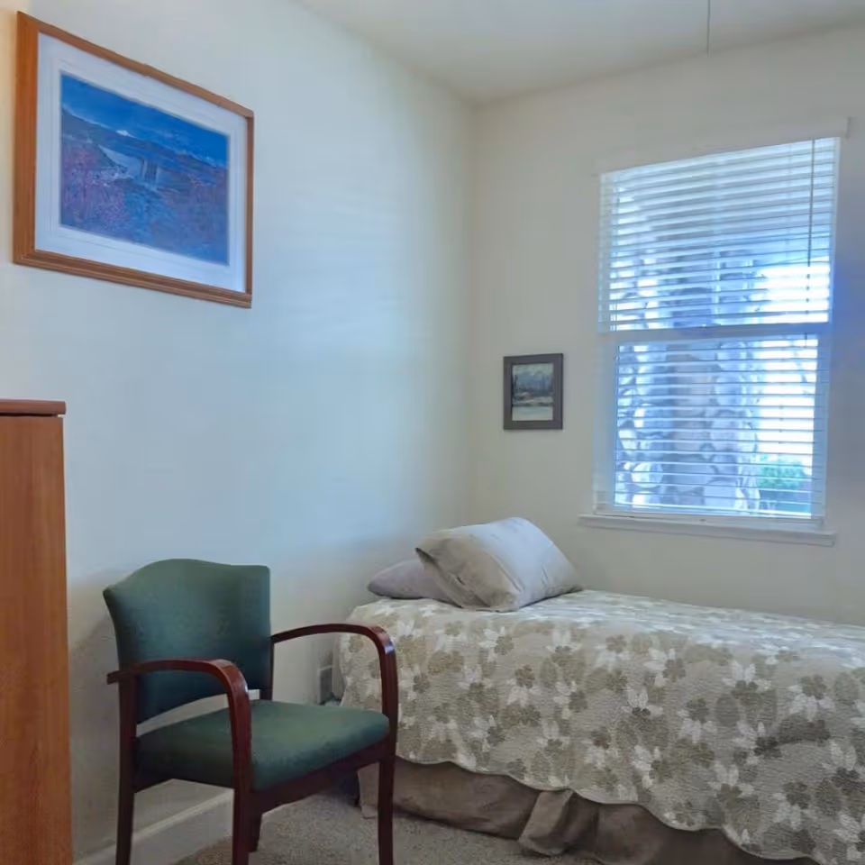 A small bedroom with a single bed covered in a floral-patterned bedspread and two pillows. Next to the bed is a green upholstered chair with wooden armrests. On the wall above the chair is a framed landscape painting, and another smaller framed picture is hung near a window with white blinds partially open, letting in natural light.
