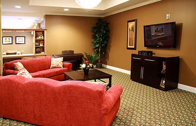 Cozy communal living room with red sofas, a coffee table, wall-mounted TV and decorative plants.