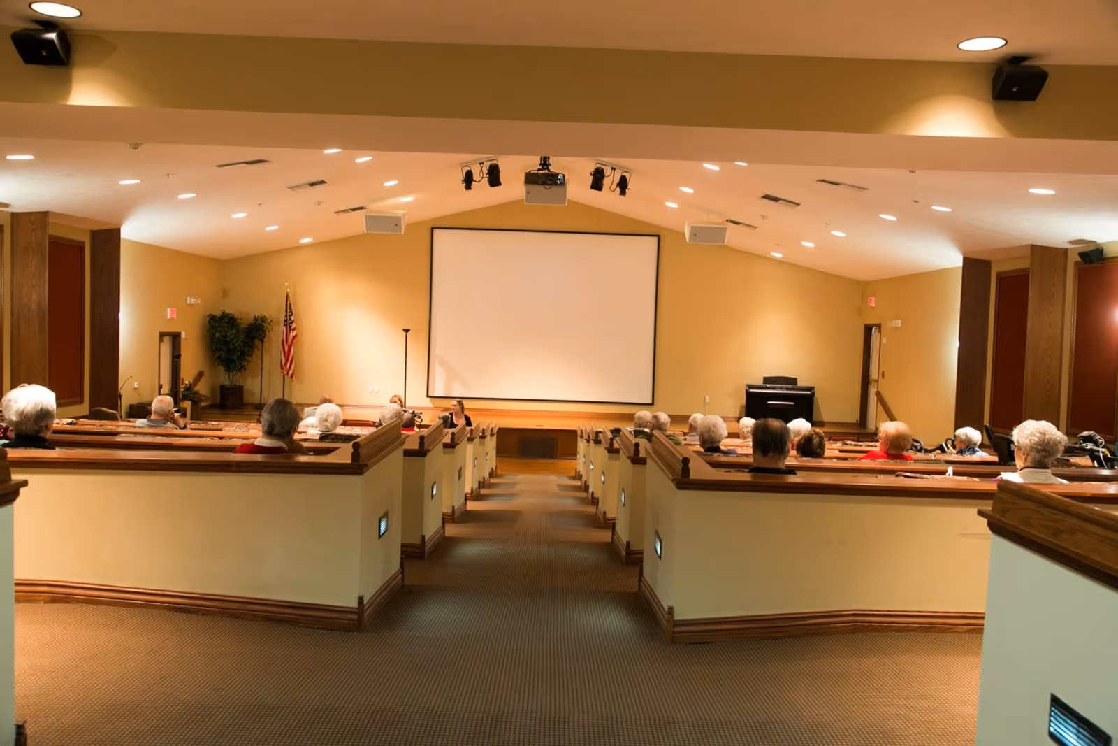 Interior view of a small auditorium or lecture hall with rows of seating occupied by elderly people facing a large blank projection screen at the front. The room has beige walls, carpeted floor, and ceiling-mounted lights and speakers. An American flag and a piano are visible near the screen.