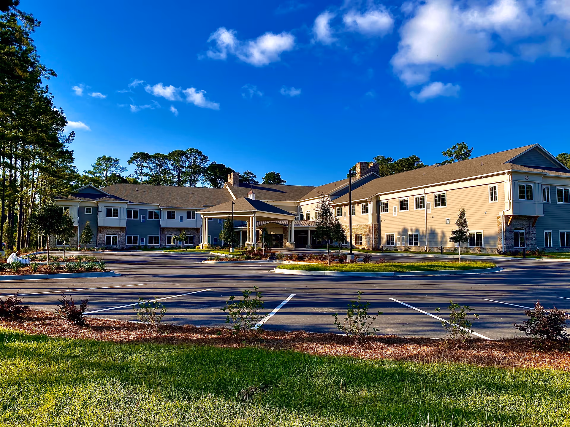 Front exterior of a two-story senior living building with a porte-cochère, parking lot, landscaping, and a bright blue sky.
