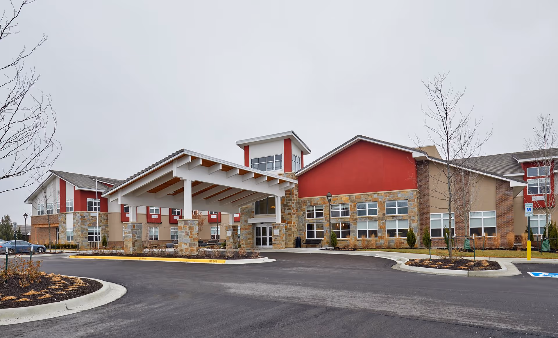 Exterior view of Leawood nursing home building with a covered entrance, stone and red siding, multiple windows, and a paved driveway with landscaped areas and leafless trees.