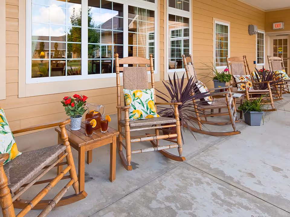 A row of wooden rocking chairs with colorful cushions on a covered porch outside a building. There are potted plants and a small wooden table with a pitcher and two glasses of iced tea with lemon slices. The building has beige siding and large windows reflecting the outdoor scenery.