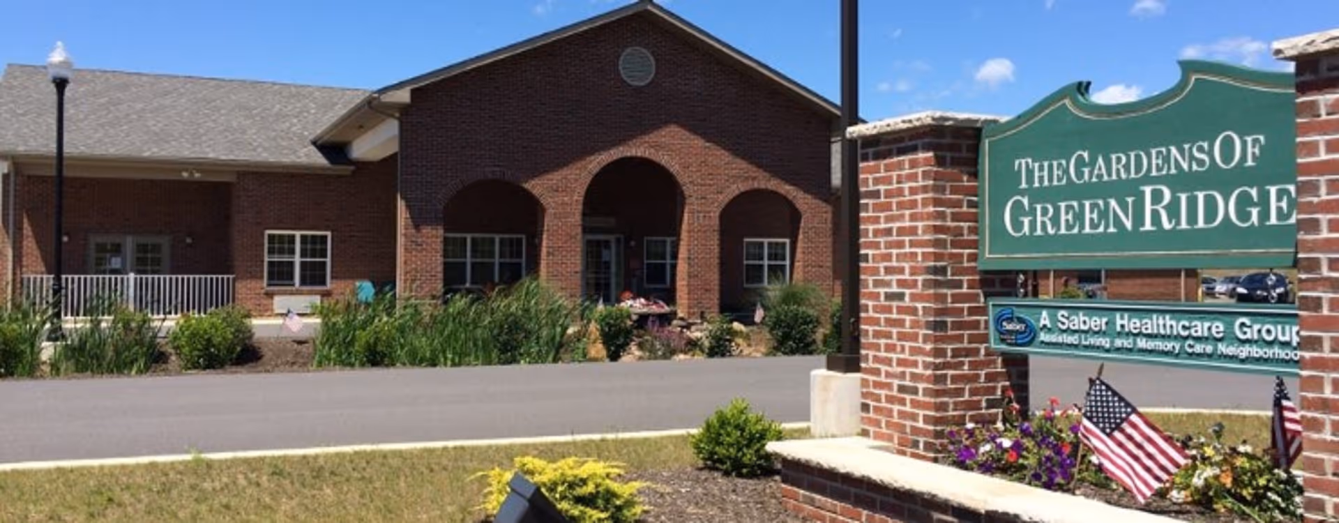 Exterior view of The Gardens of Green Ridge senior living facility showing a brick building with arched entryways, windows, and a landscaped area with plants and flowers. A green sign with white text displays the facility name along with a smaller sign indicating it is part of A Saber Healthcare Group. Two American flags are planted near the sign.