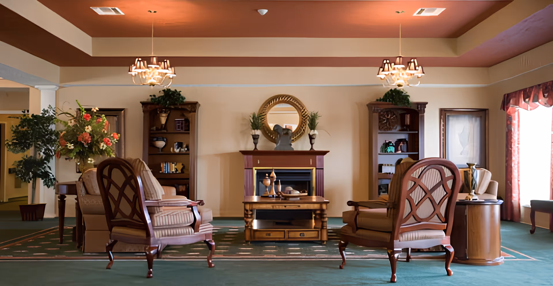 A cozy living room area with two wooden armchairs with striped cushions facing a wooden coffee table and a fireplace. The fireplace has decorative vases and a round mirror above it. There are two wooden bookshelves flanking the fireplace, each with plants and decorative items. The room has a green carpet, a reddish ceiling, and two chandeliers hanging from the ceiling. A large floral arrangement and a potted plant are visible on the left side, and a window with red curtains is on the right.