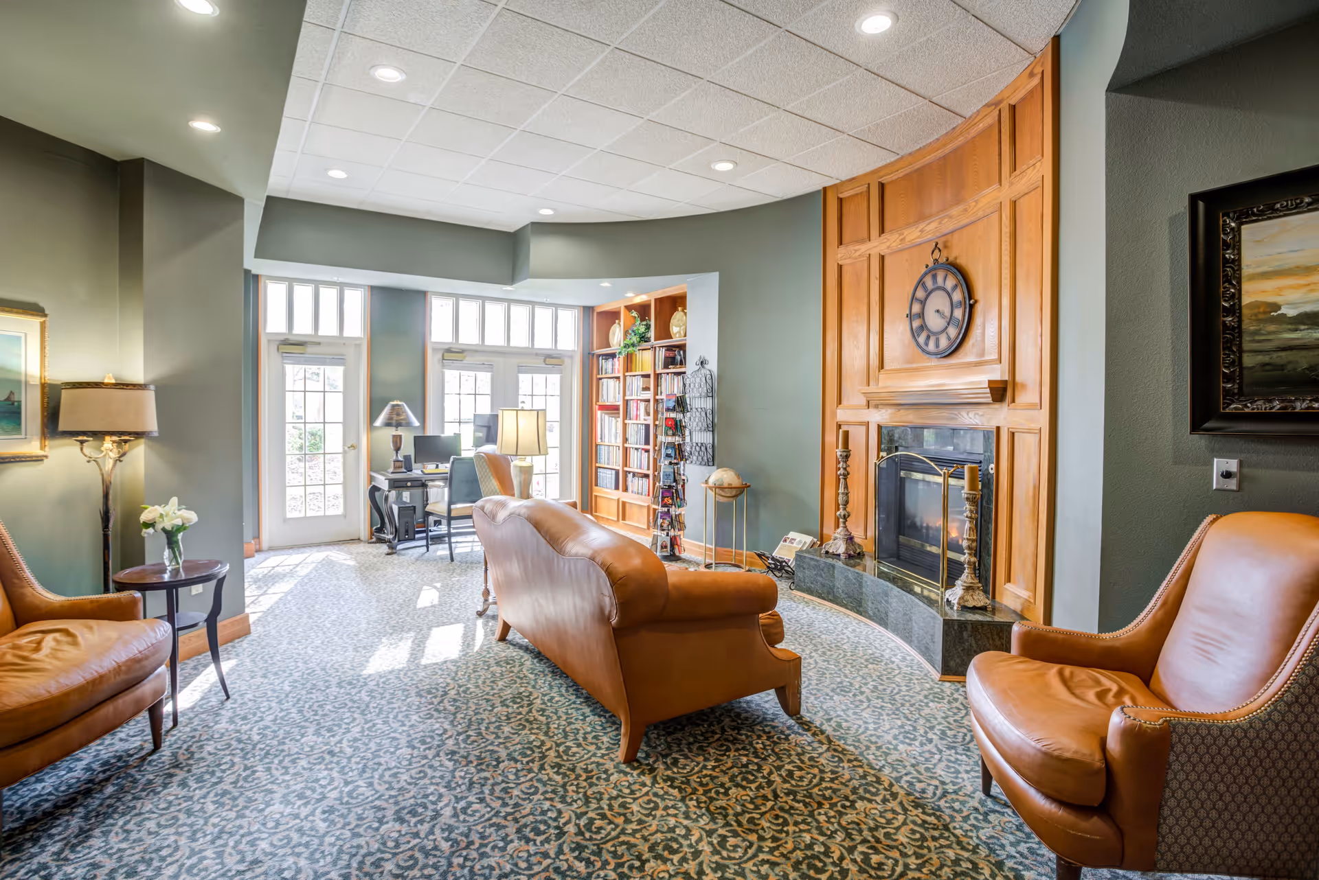 A cozy living room area with a patterned carpet, leather armchairs, and a leather sofa facing a fireplace with a wooden mantel and a large clock above it. There is a bookshelf filled with books and decorative items, a globe on a stand, and two desks with chairs near glass doors letting in natural light.