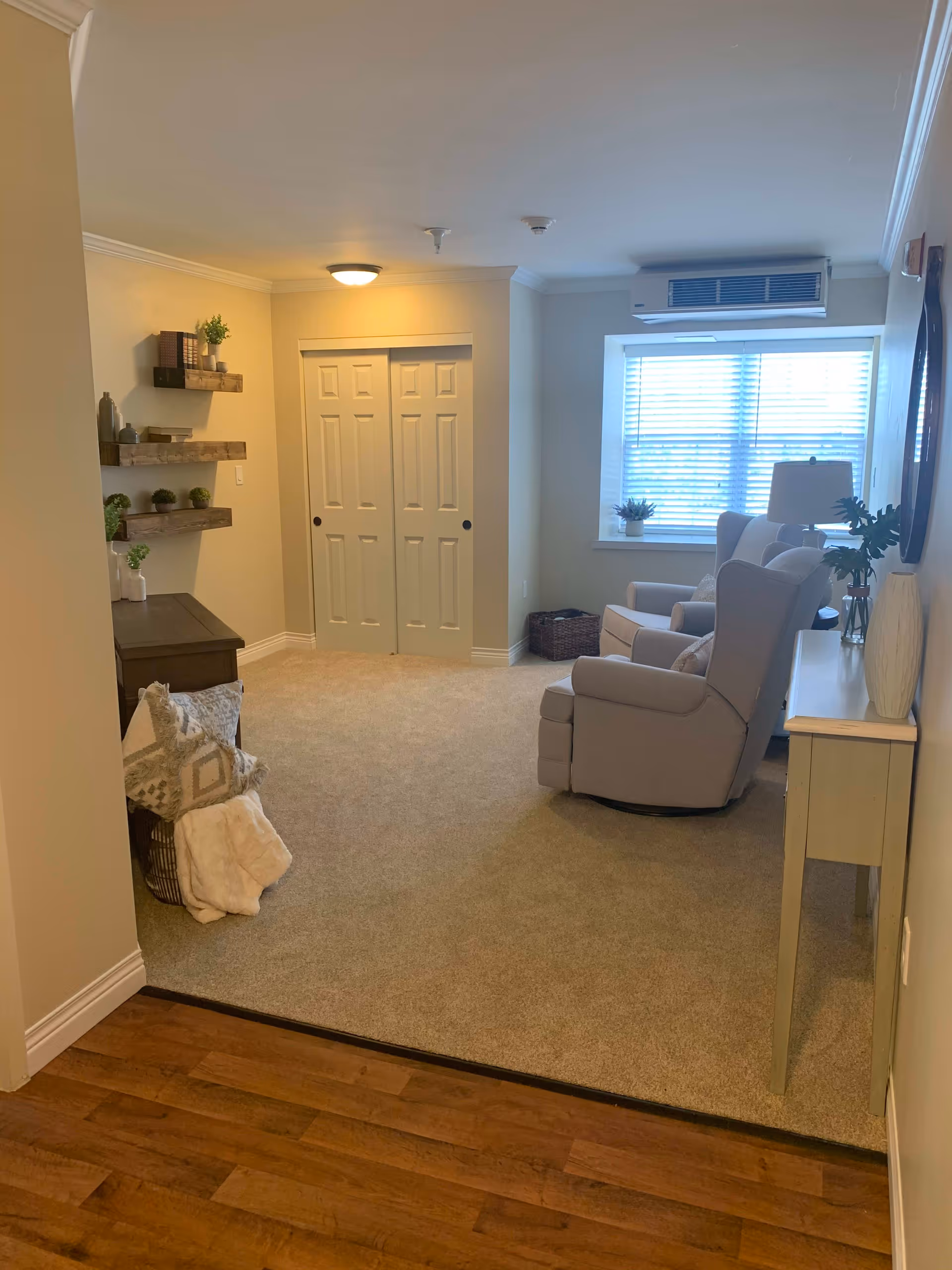 A cozy living room area in a senior living facility with beige carpet and light-colored walls. The room features two cushioned armchairs near a window with blinds, a small side table with a lamp and plant, and a console table with decorative items. There are wooden floating shelves on the left wall with small plants and books. The floor transitions from wood to carpet, and there is a closed double-door closet at the back.