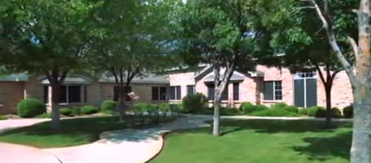 Exterior view of a brick building with multiple windows, surrounded by green grass, trees, and bushes, with a curved concrete walkway in the foreground.