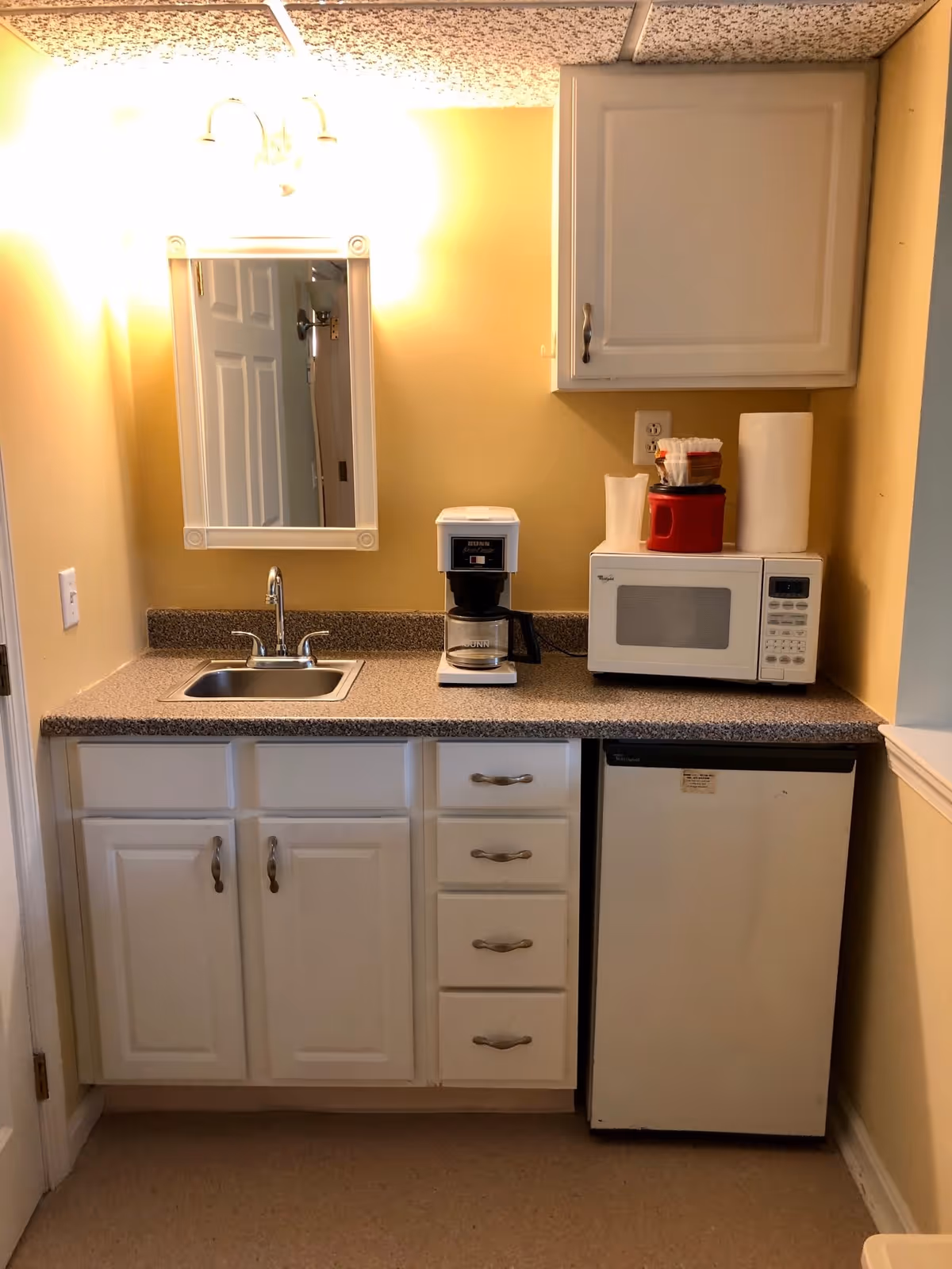 A small kitchenette area with a countertop, a sink with a faucet, a coffee maker, a microwave, and a mini refrigerator. There are white cabinets below and above the countertop. On the countertop next to the microwave are paper towels and a red container. A mirror is mounted on the wall above the sink, reflecting a door and part of the room behind.