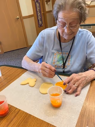 An elderly woman wearing glasses and a light blue shirt is sitting at a wooden table, painting cookies with orange icing using a small brush. There are two unpainted cookies and a cup of orange icing on the table in front of her.