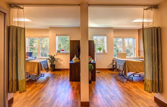 A bright and clean rehabilitation room with two hospital beds separated by a central divider. Each bed is positioned near large windows letting in natural light. The room has wooden flooring, privacy curtains on either side, and small plants placed on windowsills and near the beds.